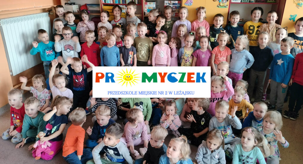 Grupa dzieci przedszkolnych w sali stojących i siedzących pozujących do zdjęcia grupowego. W centrum logo przedszkola.