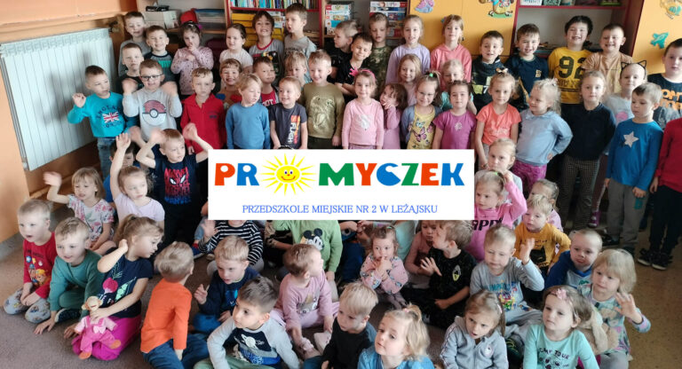 Grupa dzieci przedszkolnych w sali stojących i siedzących pozujących do zdjęcia grupowego. W centrum logo przedszkola.
